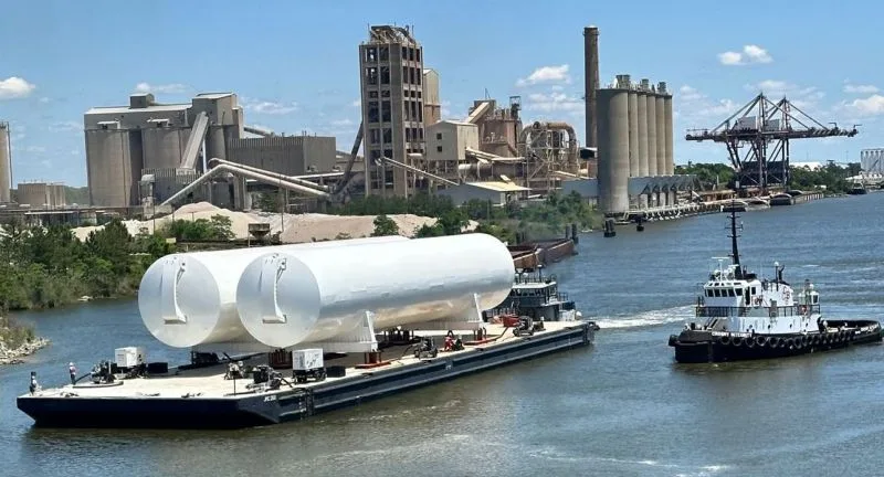 Two finished white 450,000-gallon cryogenic tanks riding a barge past an industrial waterfront, a tugboat alongside